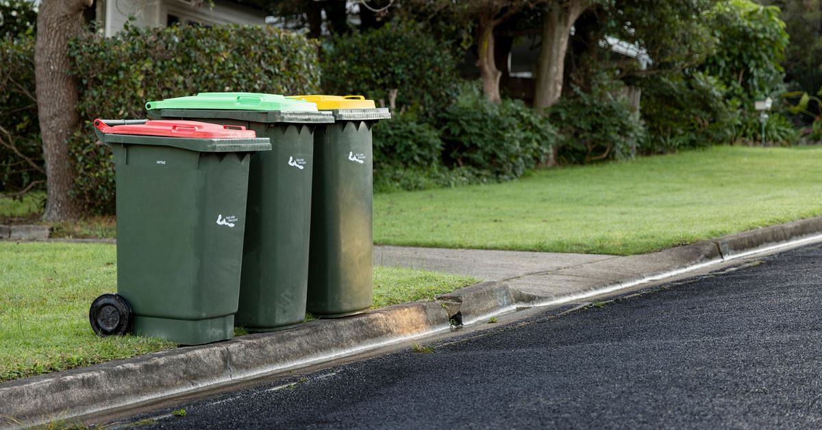 Three plastic wheelie bins lined up on a nature strip. One has a green lid, the other a yellow lid and the last a red lid
