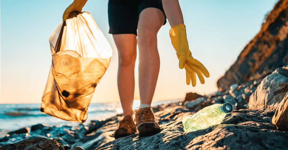 Someone bending to pick up an empty plastic bottle on a beach. The person is wearing rubber gloves and carrying a plastic bag.