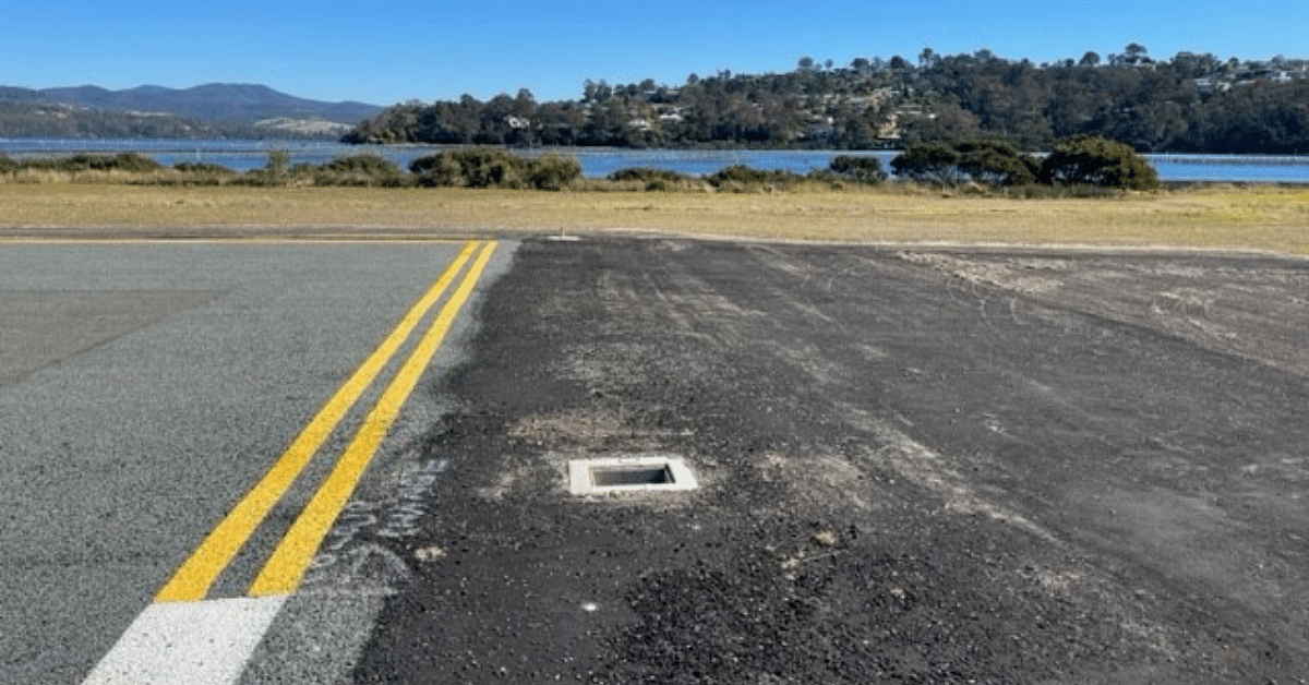 Merimbula runway surface showing a square hole where a light used to be. Merimbula lake and mountains in the background.