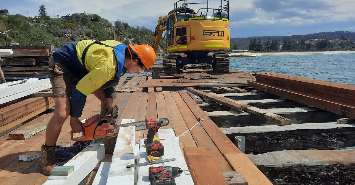 A man using a chainsaw to cut thick beams of wood while standing on the partially restored deck of the Tathra Wharf