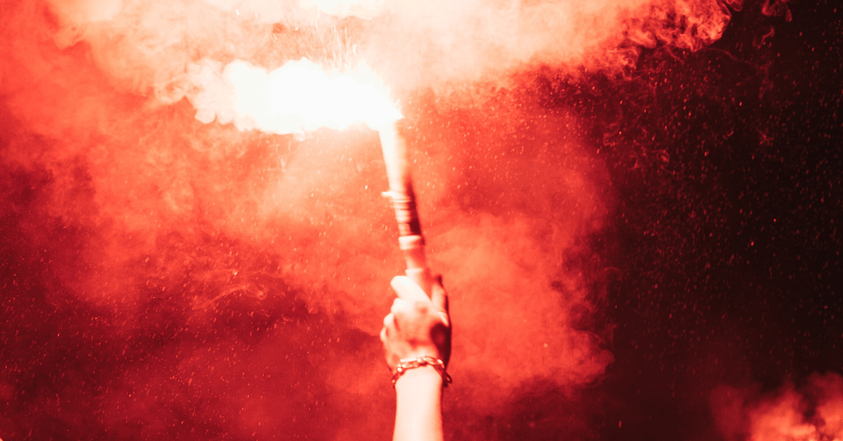 Image of a hand holding up an active marine distress flare, filling the frame with red smoke