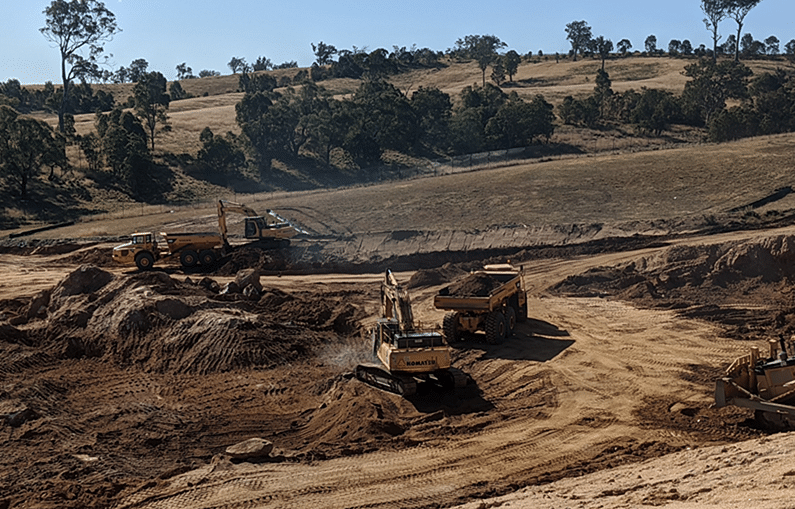 Earthmoving trucks and equipment moving soil against the backdrop of a hill studded with trees